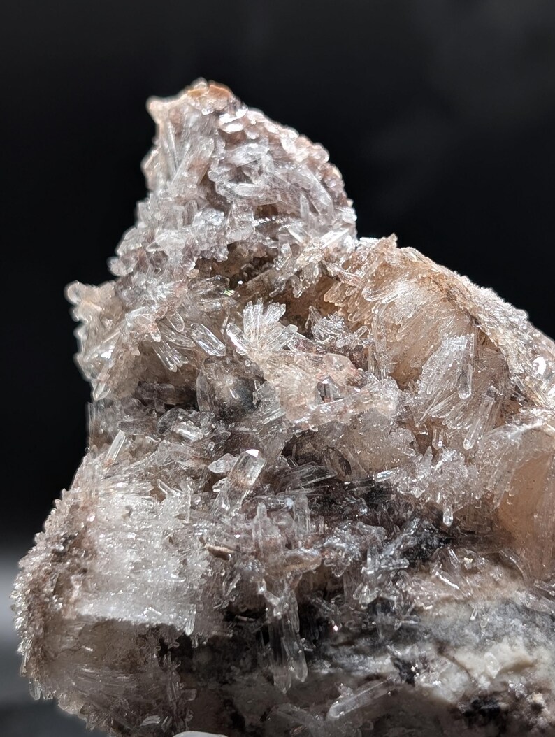May include: Close-up of a mineral specimen featuring clusters of transparent, crystal-like formations. The crystals are a clear, icy color, with hints of brown and tan. The base of the specimen is a mix of gray and brown tones, creating a natural, earthy appearance.