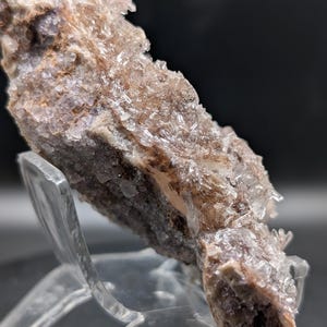 May include: A close-up of a mineral specimen with a crystalline structure. The mineral features a mix of colors, including shades of brown, purple, and clear crystals. The specimen is displayed on a clear acrylic stand against a dark background.