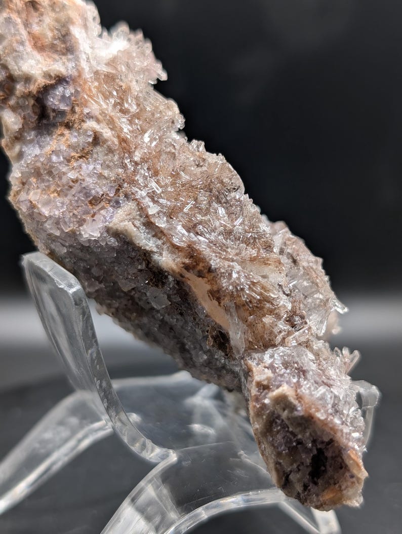 May include: A close-up of a mineral specimen with a crystalline structure. The mineral features a mix of colors, including shades of brown, purple, and clear crystals. The specimen is displayed on a clear acrylic stand against a dark background.