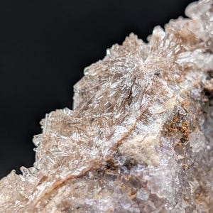 May include: Close-up of a mineral formation with crystalline structures. The crystals are translucent and appear to be white and clear, with a brown and purple base. The background is black.