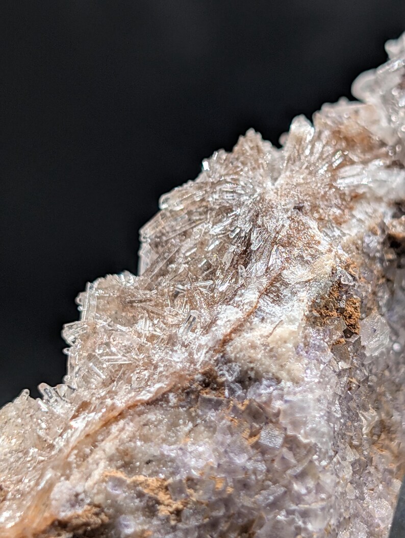 May include: Close-up of a mineral formation with crystalline structures. The crystals are translucent and appear to be white and clear, with a brown and purple base. The background is black.