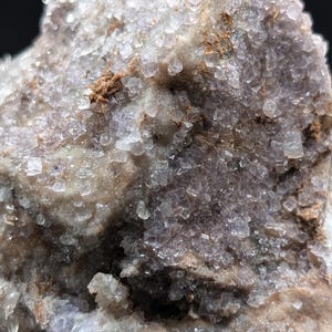 May include: Close-up of a natural mineral specimen. The rock has a crystalline structure with a mix of colors, including shades of purple, white, and brown. The surface is textured with small, cube-shaped crystals.