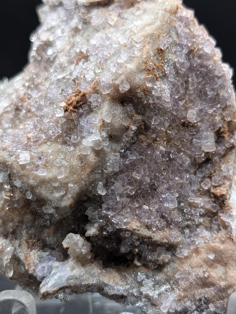 May include: Close-up of a natural mineral specimen. The rock has a crystalline structure with a mix of colors, including shades of purple, white, and brown. The surface is textured with small, cube-shaped crystals.