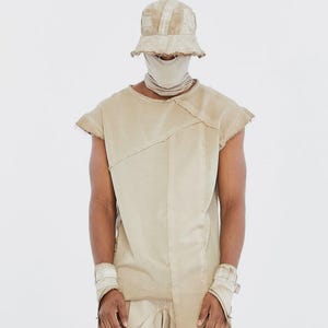 May include: A beige sleeveless top with an asymmetrical panel design and raw edges, paired with a matching bucket hat, a neck gaiter, and wrist cuffs. The ensemble features a monochromatic, weathered fabric finish with visible stitching details.