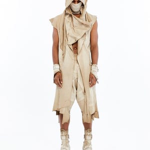 May include: A beige sleeveless hooded tunic with raw edges and a draped neckline, paired with matching loose-fitting shorts, wrist cuffs, and high-top platform boots. A fabric face covering is worn underneath the hood, completing the ensemble.