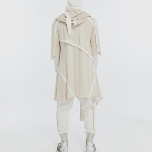 May include: A cream-colored hooded jacket with white trim and matching pants. The jacket has an asymmetrical design and short sleeves. The pants have a zipper detail at the ankles. The outfit is paired with silver and gray boots.