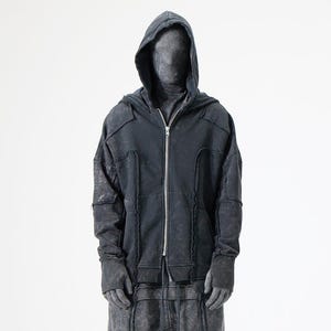 Può includere: Una felpa con cappuccio e zip color grigio antracite, caratterizzata da una texture slavata e vissuta con cuciture a vista e bordi grezzi. Il capo presenta un cappuccio, maniche lunghe con polsini a coste e una chiusura a zip in metallo argentato.