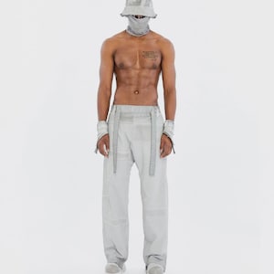 Può includere: Un cappello a secchiello grigio chiaro, una copertura per il viso abbinata, polsini e pantaloni larghi con costruzione a pannelli. L'insieme presenta una tonalità grigia uniforme e tenue, con lunghe strisce di tessuto in vita.