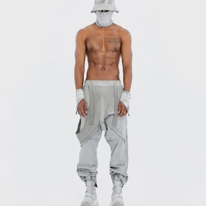 May include: A person wearing a light gray outfit, including a bucket hat, face covering, and pants with attached apron-like fabric. The outfit is paired with matching wrist cuffs and chunky boots. The background is plain white.