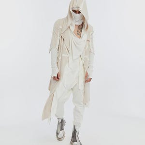 May include: A person wearing a layered cream-colored outfit with a hooded jacket, draped top, and white pants. The outfit includes a face covering and silver-grey boots with thick soles. The overall style is avant-garde and modern.