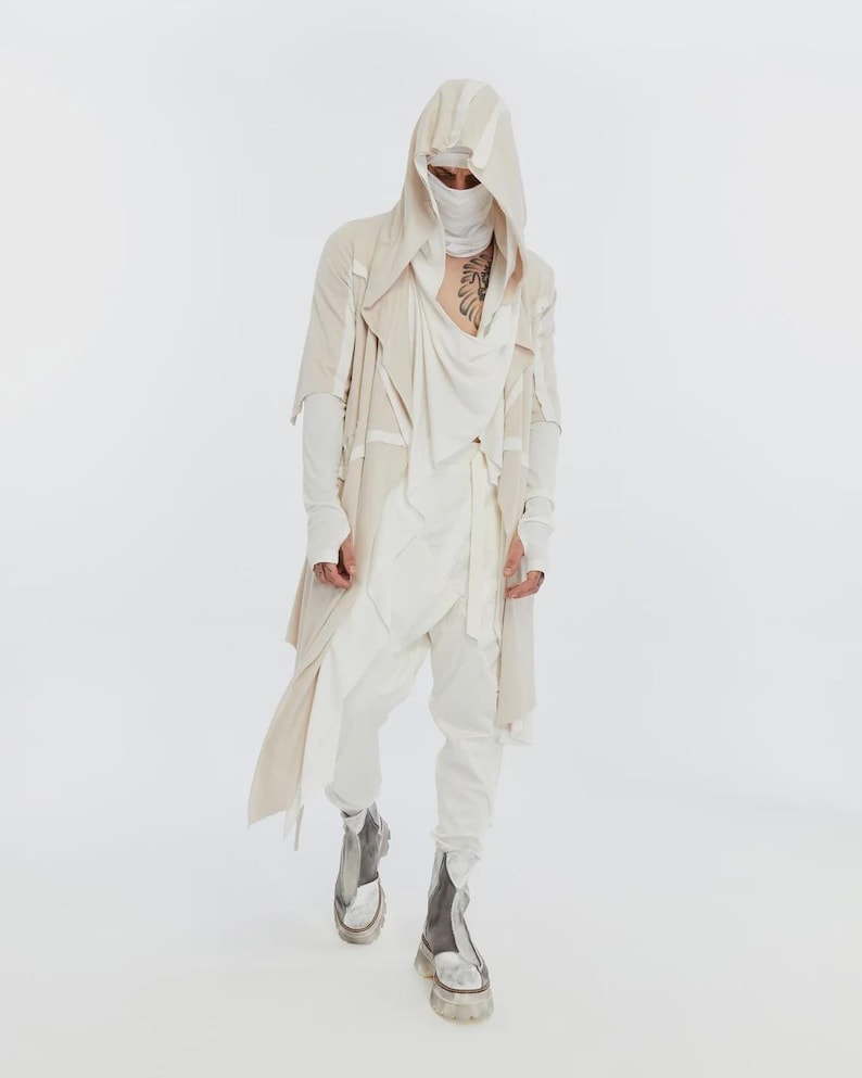 May include: A person wearing a layered cream-colored outfit with a hooded jacket, draped top, and white pants. The outfit includes a face covering and silver-grey boots with thick soles. The overall style is avant-garde and modern.