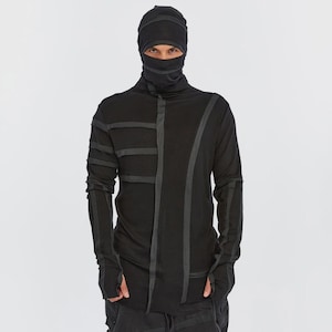 Op de afbeelding: Een zwart shirt met lange mouwen, een hoge kraag, een geïntegreerde capuchon en een gezichtsbedekking. Het kledingstuk heeft donkergrijze verticale en horizontale stroken stof op de borst en mouwen, met duimgaten bij de manchetten.