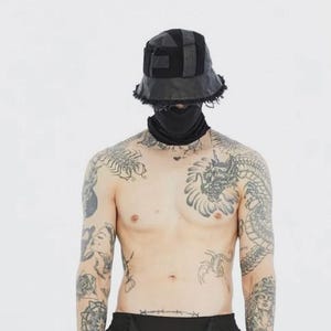 Puede incluir: Sombrero de pescador negro con diseño de retazos y bordes deshilachados. El sombrero se usa con una cubierta facial negra. La persona de la imagen tiene múltiples tatuajes en brazos y pecho.