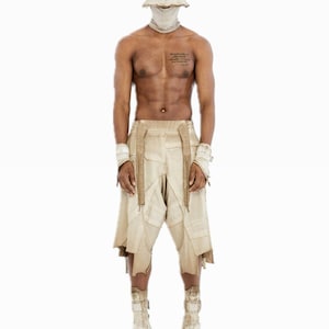 May include: A person wearing a beige outfit, including a bucket hat, face covering, shorts, wristbands, and platform boots. The shorts have a patchwork design. The outfit is a neutral color.