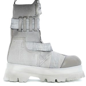 May include: A high-top boot in shades of light gray and white, featuring a ribbed knit upper, multiple horizontal fabric straps with metal hardware, a reinforced rounded toe cap, and a thick, chunky white lug sole with deep tread patterns.