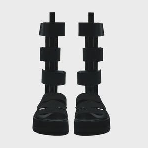 May include: Pair of black platform sandals with multiple wide straps and a high shaft. The sandals have a chunky sole and are designed with a series of vertical straps extending up the leg. The footwear is displayed against a plain background.