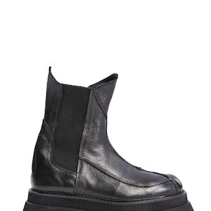 May include: A black ankle boot with a textured leather upper, a high shaft, and a wide elastic side panel. The boot features a prominent vertical seam down the front, a rounded toe, and a thick, chunky black lug sole with deep tread patterns.