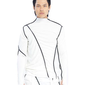 Può includere: Una maglia a maniche lunghe con collo alto color bianco sporco, caratterizzata da evidenti cuciture nere a vista che creano un motivo geometrico asimmetrico su petto, spalle e maniche, con fori per i pollici visibili sui polsini.