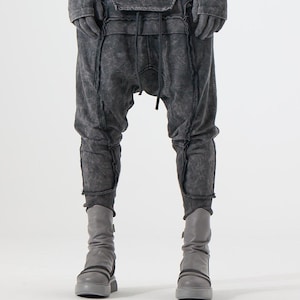 Peut inclure: Pantalon à entrejambe bas gris anthracite foncé avec une texture délavée et usée, présentant des coutures apparentes, une taille à cordon de serrage et un haut à manches longues assorti. Bottes mi-mollet gris clair à semelles épaisses.