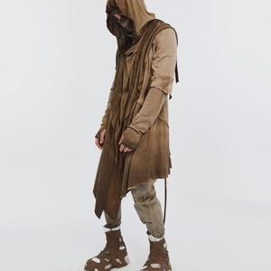 May include: A full-length shot of a person wearing a layered brown hooded cloak and matching pants. The outfit includes a face covering, long sleeves, and unique brown and white cutout shoes. The overall style suggests a post-apocalyptic or dystopian theme.