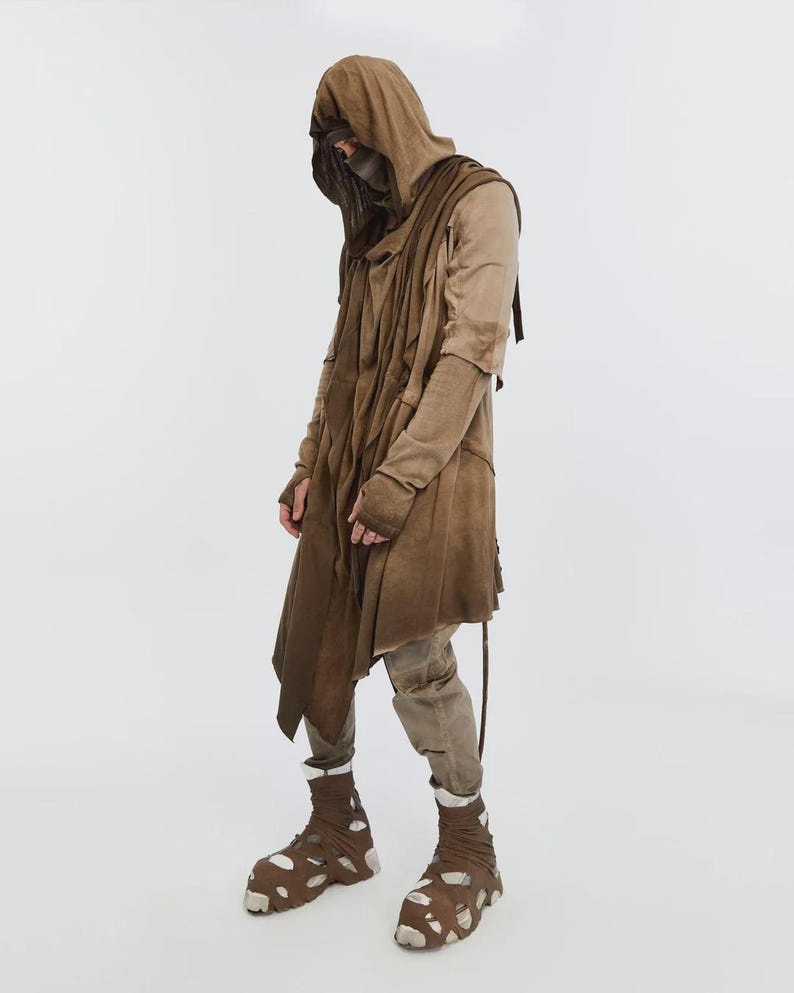 May include: A full-length shot of a person wearing a layered brown hooded cloak and matching pants. The outfit includes a face covering, long sleeves, and unique brown and white cutout shoes. The overall style suggests a post-apocalyptic or dystopian theme.