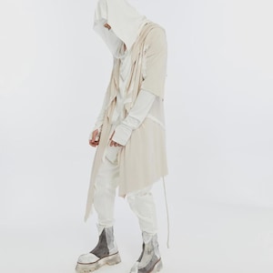 May include: A person wearing a layered outfit in shades of white and beige. The ensemble includes a hooded top, long-sleeved shirt, draped vest, and matching pants. The look is completed with chunky, two-tone boots with clear soles.