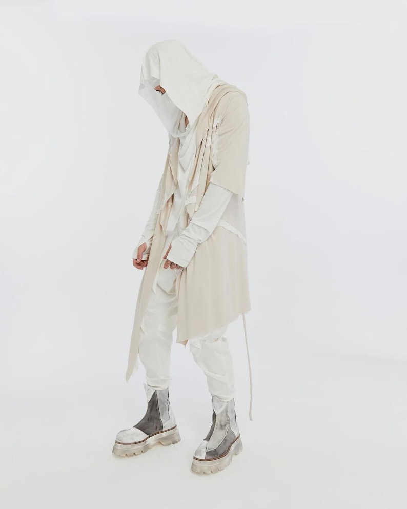 May include: A person wearing a layered outfit in shades of white and beige. The ensemble includes a hooded top, long-sleeved shirt, draped vest, and matching pants. The look is completed with chunky, two-tone boots with clear soles.