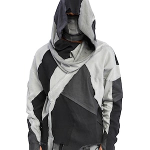 Puede incluir: Una sudadera con capucha que presenta un diseño de patchwork con paneles de tela negra y gris con bordes crudos y deshilachados. La prenda incluye una cubierta facial integrada que se extiende desde el cuello, mangas largas y corte holgado.
