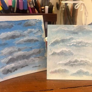 Puede incluir: Dos pinturas cuadradas con estudios de nubes. La pintura de la izquierda muestra nubes de color gris oscuro sobre un cielo azul vibrante. La pintura de la derecha presenta nubes más suaves de color gris claro y blanco sobre un cielo azul pálido.