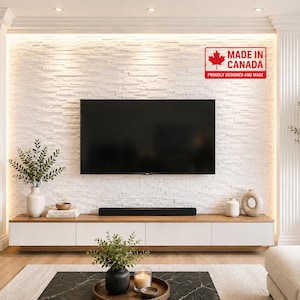 May include: A wall-mounted television centered on a white textured stone wall with integrated lighting, positioned above a long white and wood-toned media console. A red and white label reads MADE IN CANADA PROUDLY DESIGNED AND MADE.