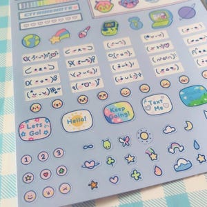 May include: A sheet of light blue stickers with various designs. The stickers include text like "Hello!", "Keep Going!", and "Text Me". There are also illustrations of planets, stars, clouds, and smiley faces. The sheet is on a blue and white checkered surface.