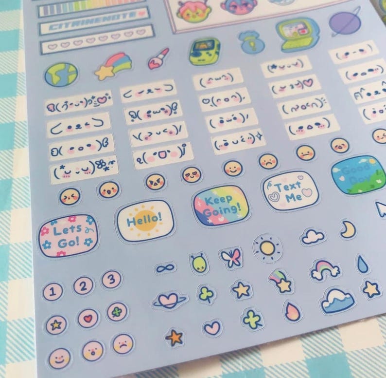 May include: A sheet of light blue stickers with various designs. The stickers include text like "Hello!", "Keep Going!", and "Text Me". There are also illustrations of planets, stars, clouds, and smiley faces. The sheet is on a blue and white checkered surface.