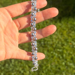 Könnte beinhalten: Ein silbernes Armband mit abwechselnden Reihen aus grauen Perlen und hellblauen Edelsteinen. Das Armband wird vor einem grünen Hintergrund gehalten, der sein zartes Design und seine funkelnden Details hervorhebt. Der Verschluss hat die Form eines dekorativen Herzens.