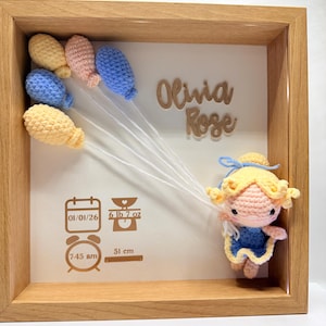 Könnte beinhalten: Ein gerahmtes Baby-Ankündigungsbild mit einer gehäkelten Puppe und Luftballons. Die Ankündigung enthält den Namen "Olivia Rose", Datum, Uhrzeit, Gewicht (3,04 kg) und Größe (51 cm). Die Ballons sind gelb, rosa und blau.