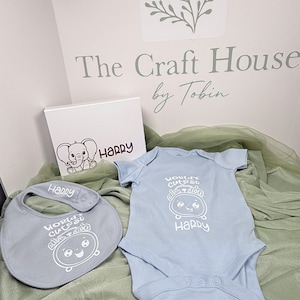 May include: A light blue baby bodysuit and matching bib featuring a white graphic of an alarm clock with the text "WORLD'S CUTEST ALARM CLOCK" and the name "HARRY" printed in white. A white box with an elephant illustration and the name "HARRY" is included.
