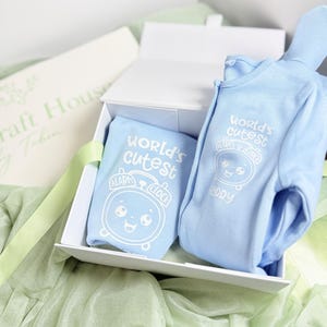 May include: A light blue baby onesie and a matching folded garment inside a white gift box. Both items feature a white graphic print of a smiling alarm clock with the text "World's cutest" above and the name "Harry" below the clock.