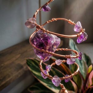 Può includere: Una scultura in filo di rame con cristalli di ametista. Il design a spirale incorpora pietre di ametista grezze e pezzi di ametista più piccoli e lucidi, creando un pezzo decorativo unico. La scultura è esposta su uno sfondo sfocato.