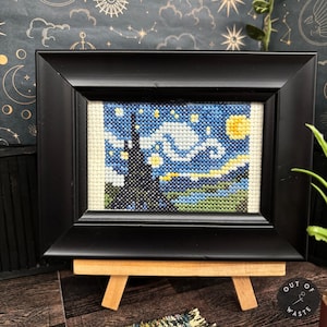 May include: A framed cross-stitch of Van Gogh's "Starry Night" in a black frame. The artwork features a blue night sky with swirling clouds, a yellow moon, and a dark, silhouetted village. The frame is displayed on a small wooden easel.