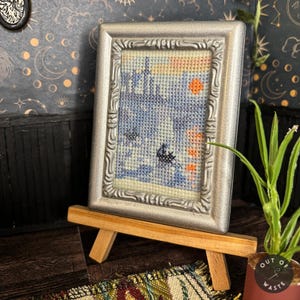 May include: A miniature framed cross-stitch artwork depicting a blue and white scene with orange accents, displayed on a small wooden easel. The silver frame adds a decorative touch to the artwork. The overall theme is miniature art.
