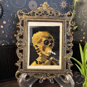 May include: A framed cross-stitch artwork featuring a yellow skull with a cigarette in its mouth. The skull is set against a black background and framed in an ornate, antique gold-colored frame. The artwork is displayed on a clear stand.