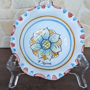 May include: A small, round, white ceramic dish with a scalloped edge featuring alternating red and blue painted dashes. The center displays a hand-painted blue and yellow flower motif with the signature F. Soliva written in black ink below it.