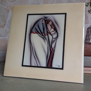 May include: A square ceramic tile featuring a centered rectangular print of two figures wrapped in blankets. The left figure wears a blue and white blanket, while the right figure wears a red and white blanket. The tile has a cream-colored border.