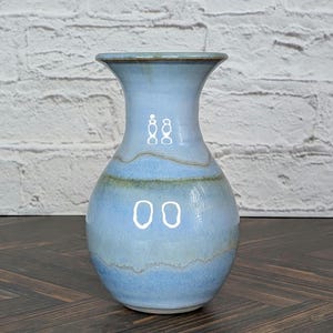 May include: A ceramic vase with a flared rim and a bulbous body, featuring a smooth, glossy glaze in shades of light blue, periwinkle, and soft tan. The surface displays horizontal bands of color with subtle variations and a reflective finish.