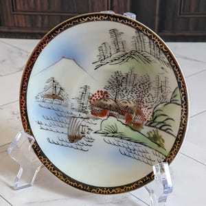 May include: A round ceramic saucer featuring a hand-painted landscape scene with a mountain, traditional houses, trees, and a boat on water. The rim is decorated with a black border containing intricate gold filigree patterns and red accents.