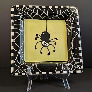 May include: A square ceramic plate featuring a central light green square with a black spider illustration hanging from a single line. The border is black with a white spiderweb pattern and a black and white checkered edge around the rim.