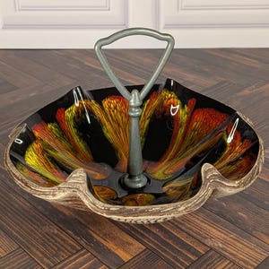 May include: A decorative serving bowl with a central metal handle. The bowl features a scalloped edge with a textured, light brown exterior and a glossy interior finish displaying a vibrant, swirling pattern of black, orange, and yellow.