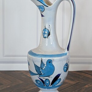 May include: A ceramic pitcher with a white glaze and hand-painted blue motifs. The design features a blue bird, butterflies, and floral elements. It has a tall, slender neck, a curved handle, and a flared base. The word Tonala is painted on the side.