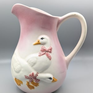 May include: A ceramic pitcher featuring a soft pink gradient finish and a raised relief design of two white geese. Each goose is adorned with a small pink and white polka-dot bow around its neck. The pitcher has a curved handle and a wide spout.