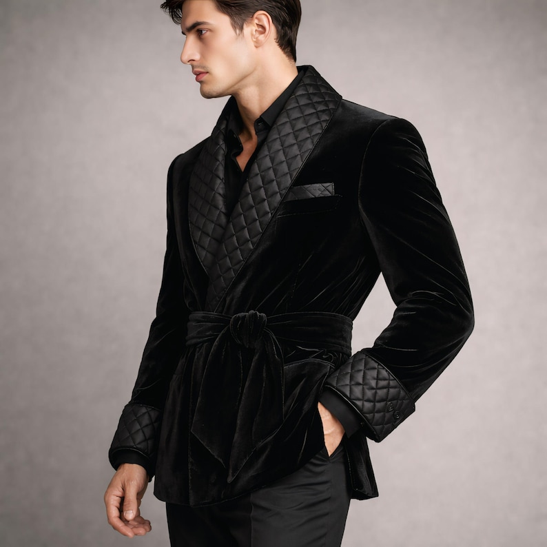 May include: A black velvet smoking jacket with a quilted shawl collar, cuffs, and pocket trim. The jacket features a matching tie belt and is worn over a black shirt and pants. The jacket is a luxurious garment.