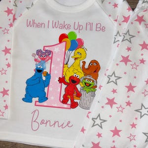 May include: White pyjama set featuring a pink number 1 and Sesame Street characters. The top has the text "When I Wake Up I'll Be" and the name "Bonnie". The pyjamas have a star pattern in pink and grey.